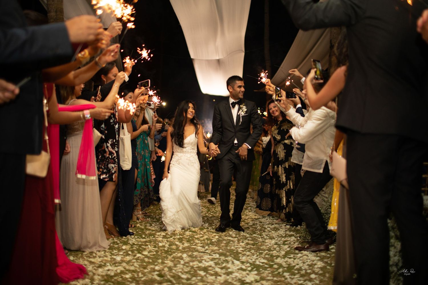 Bride and groom exit through a sparkler tunnel as confetti falls at a festive nighttime wedding reception.