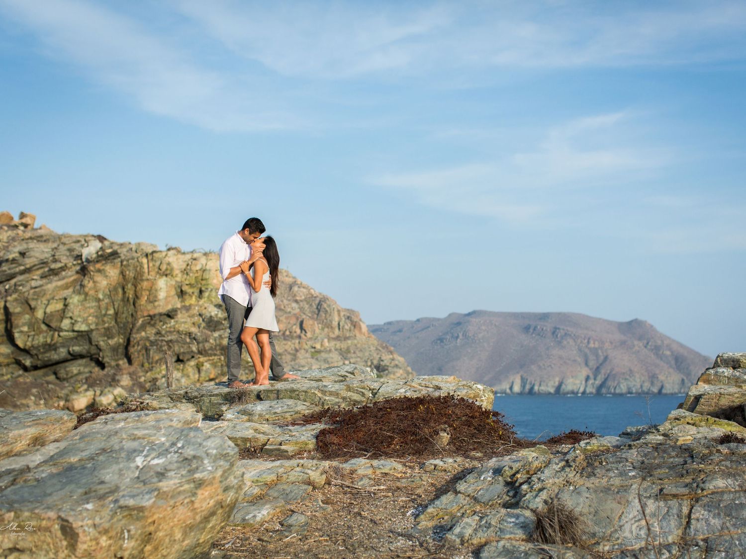 Couple kissing on rocky coastal cliffs with stunning blue ocean and mountain landscape in background.