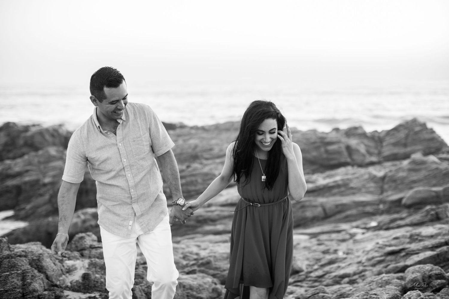 Couple holds hands walking on rocky coastal shoreline in black and white engagement photo near ocean waves.