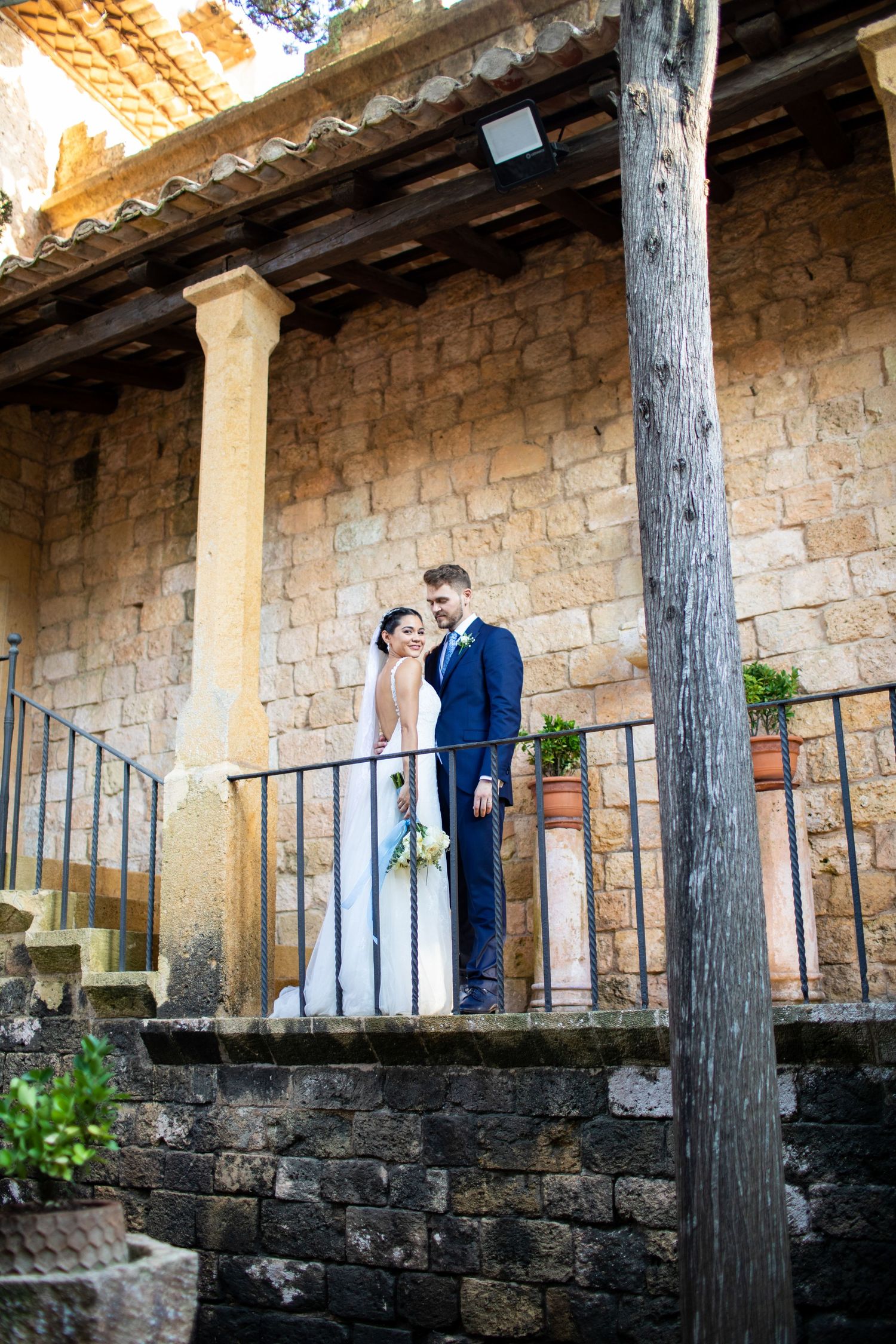 Elegant bride and groom standing on stone balcony of historic rustic venue with wooden columns.