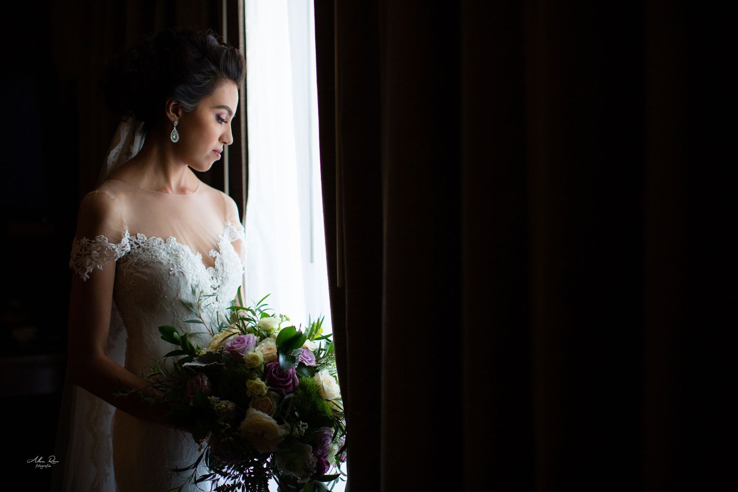 Elegant bride in lace off-shoulder gown holding colorful bouquet while gazing out a window in soft light.