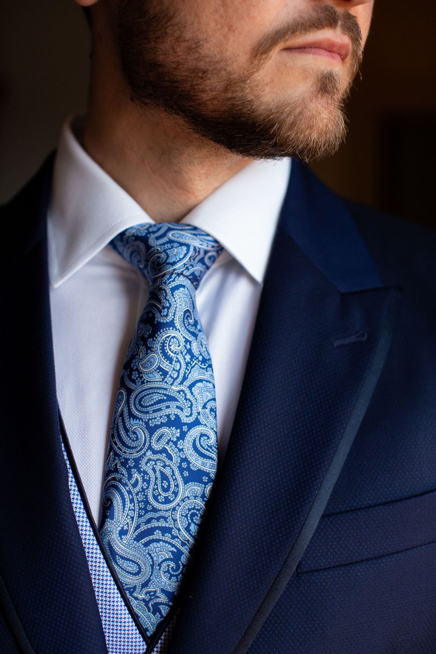 Close-up of a groom wearing a navy blue suit with an elegant blue paisley patterned tie and white dress shirt.