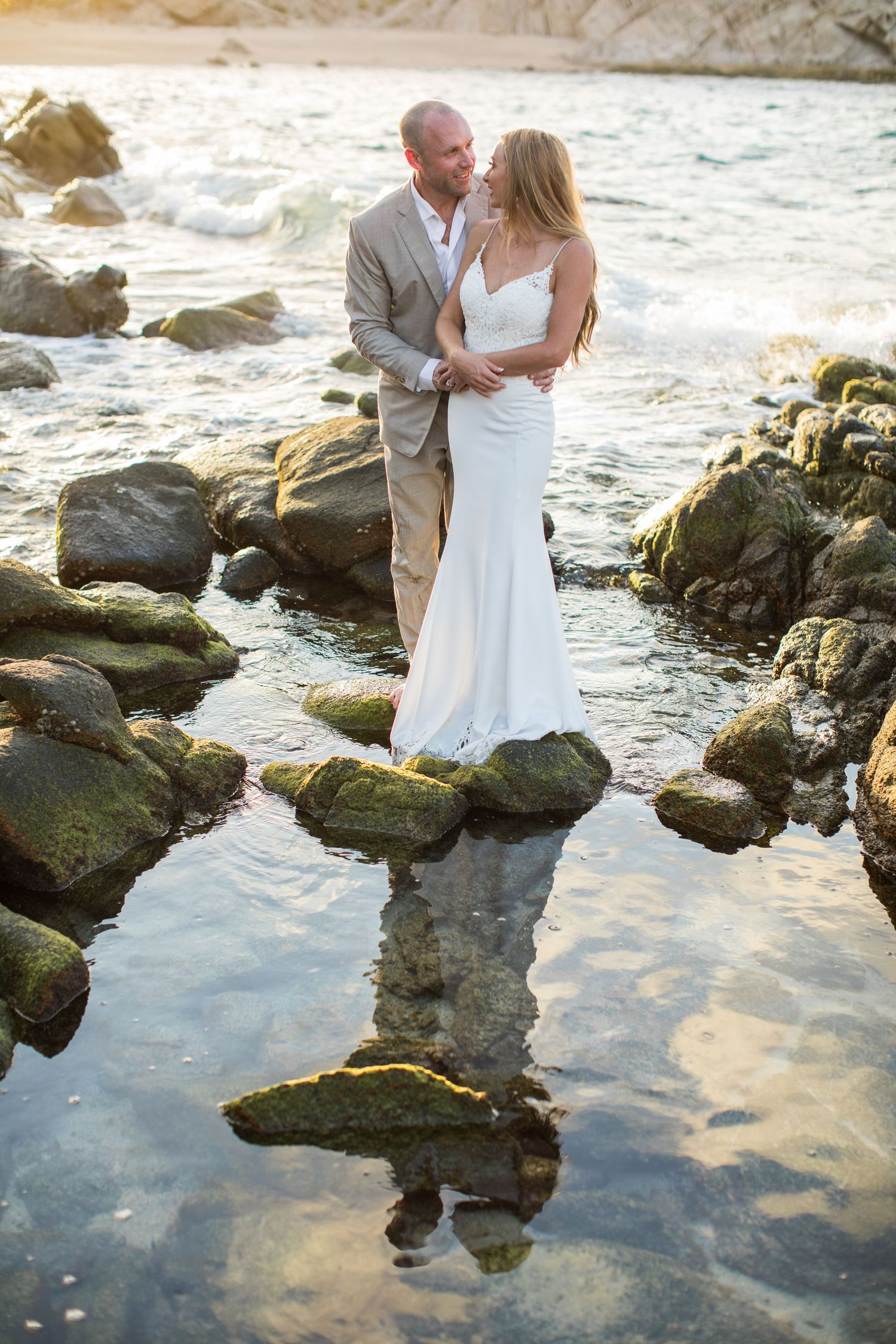Bride and groom gaze at each other on mossy rocks with their reflection in still water.