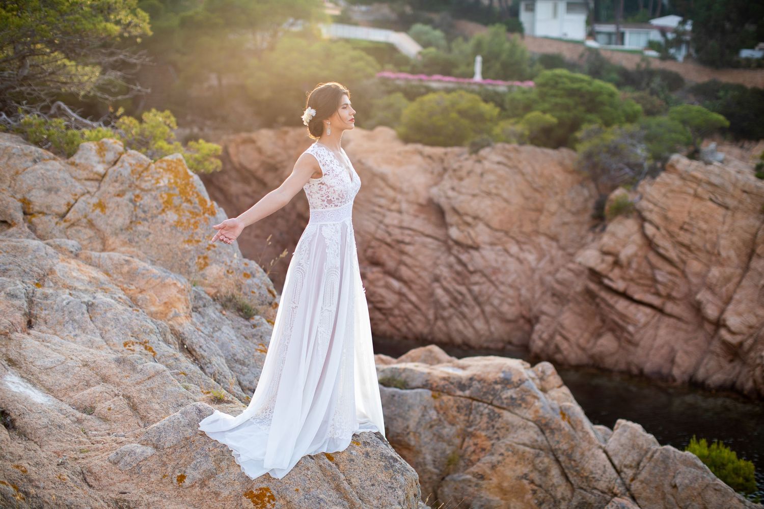 Bride in flowing white lace gown stands arms outstretched on dramatic rocky coastal cliffs at sunset.
