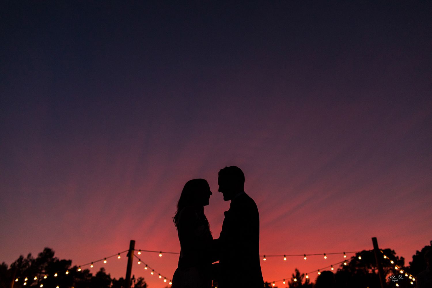 Romantic couple silhouette against vibrant red and purple sunset sky with string lights.