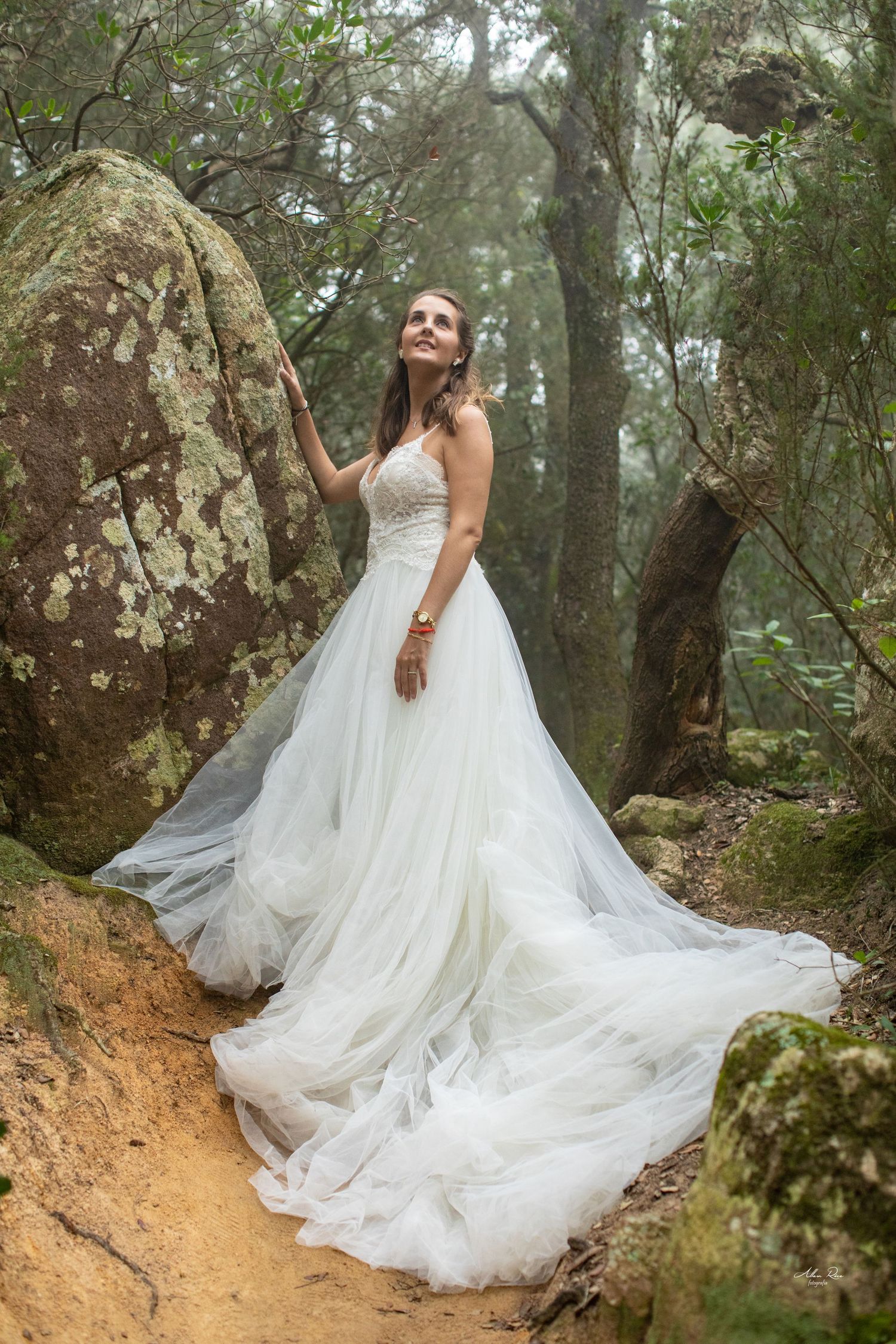 Radiant bride in strapless tulle gown poses beside mossy rocks in a misty forest.