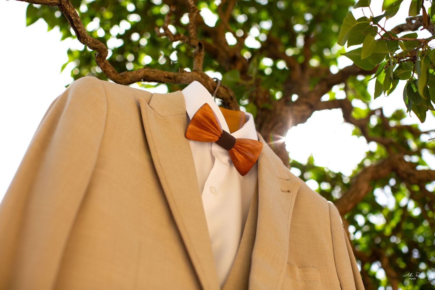Elegant beige groom suit with orange wooden bow tie hanging on a sunlit tree branch.