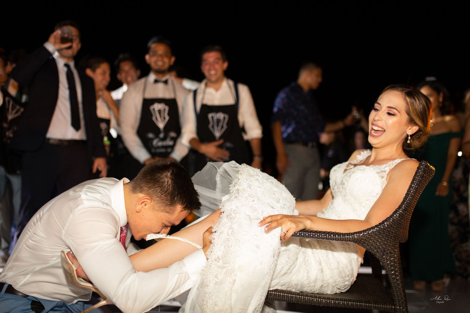 Groom playfully searching for garter under brides dress as guests cheer at reception.