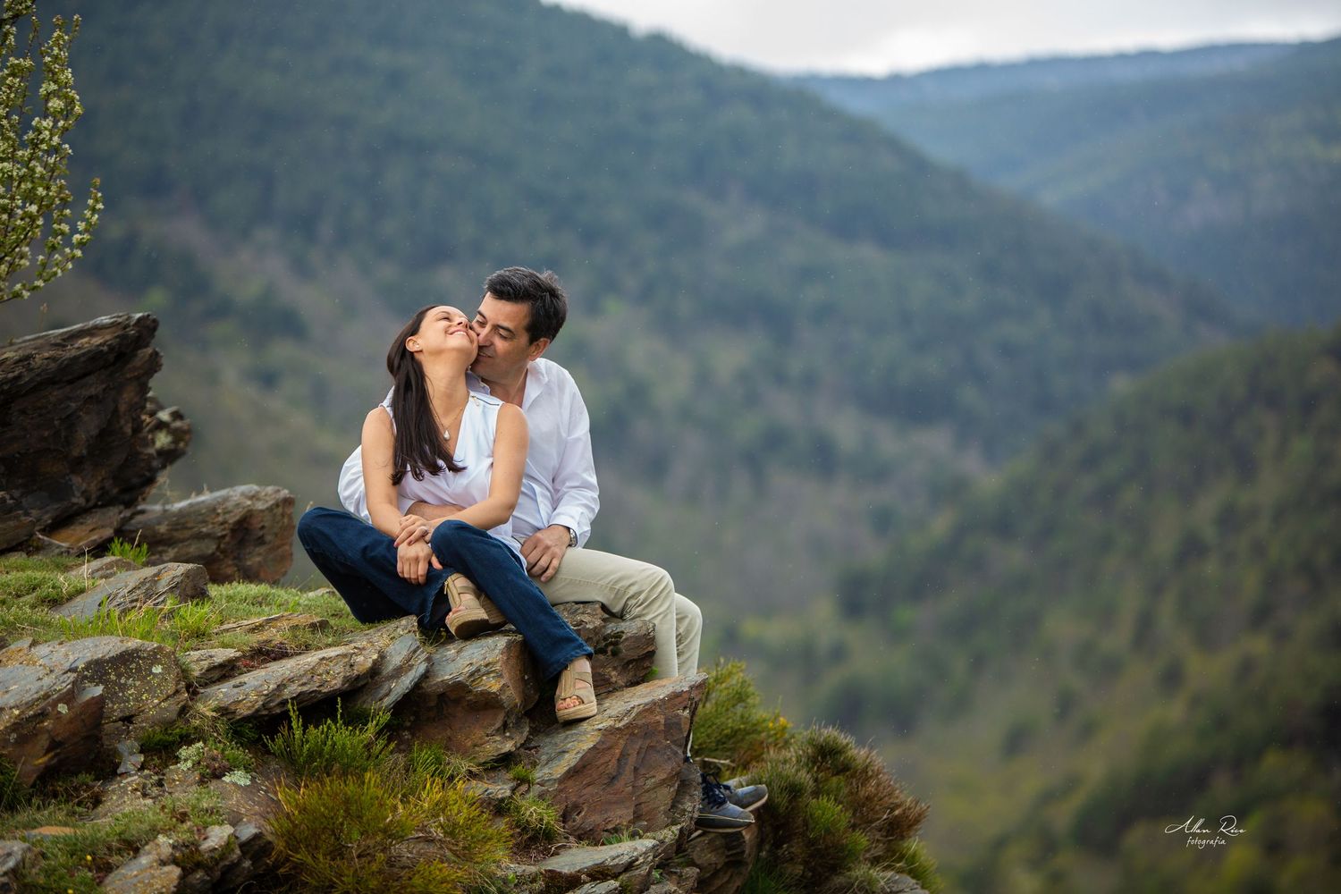 Romantic couple sitting together on rocky mountain overlook with lush green valley and forest backdrop.