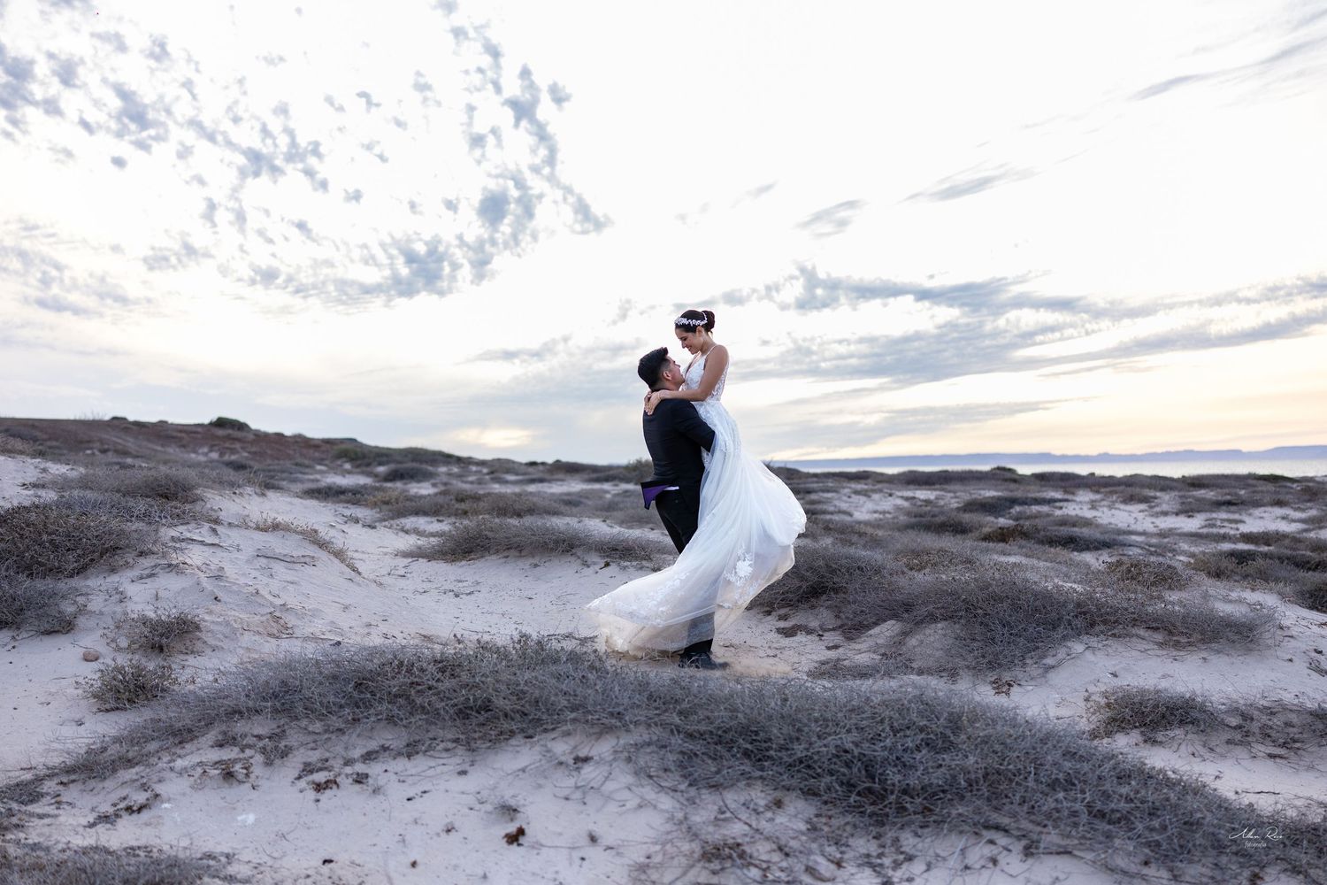 Groom dipping bride in white gown on sandy dunes under a cloudy sunset sky.
