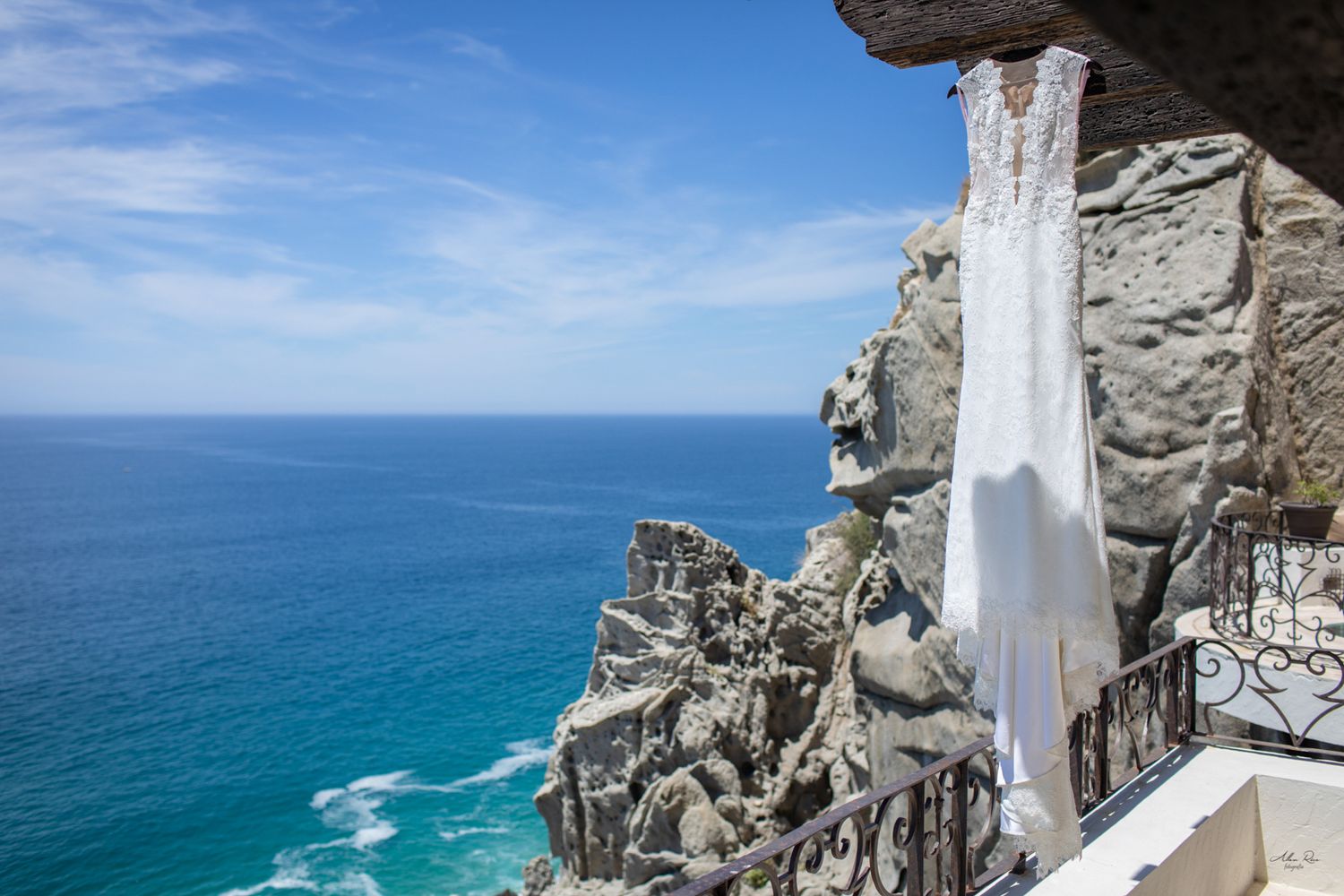 White wedding dress draped over rocky ocean cliff balcony with stunning turquoise sea and blue sky backdrop.