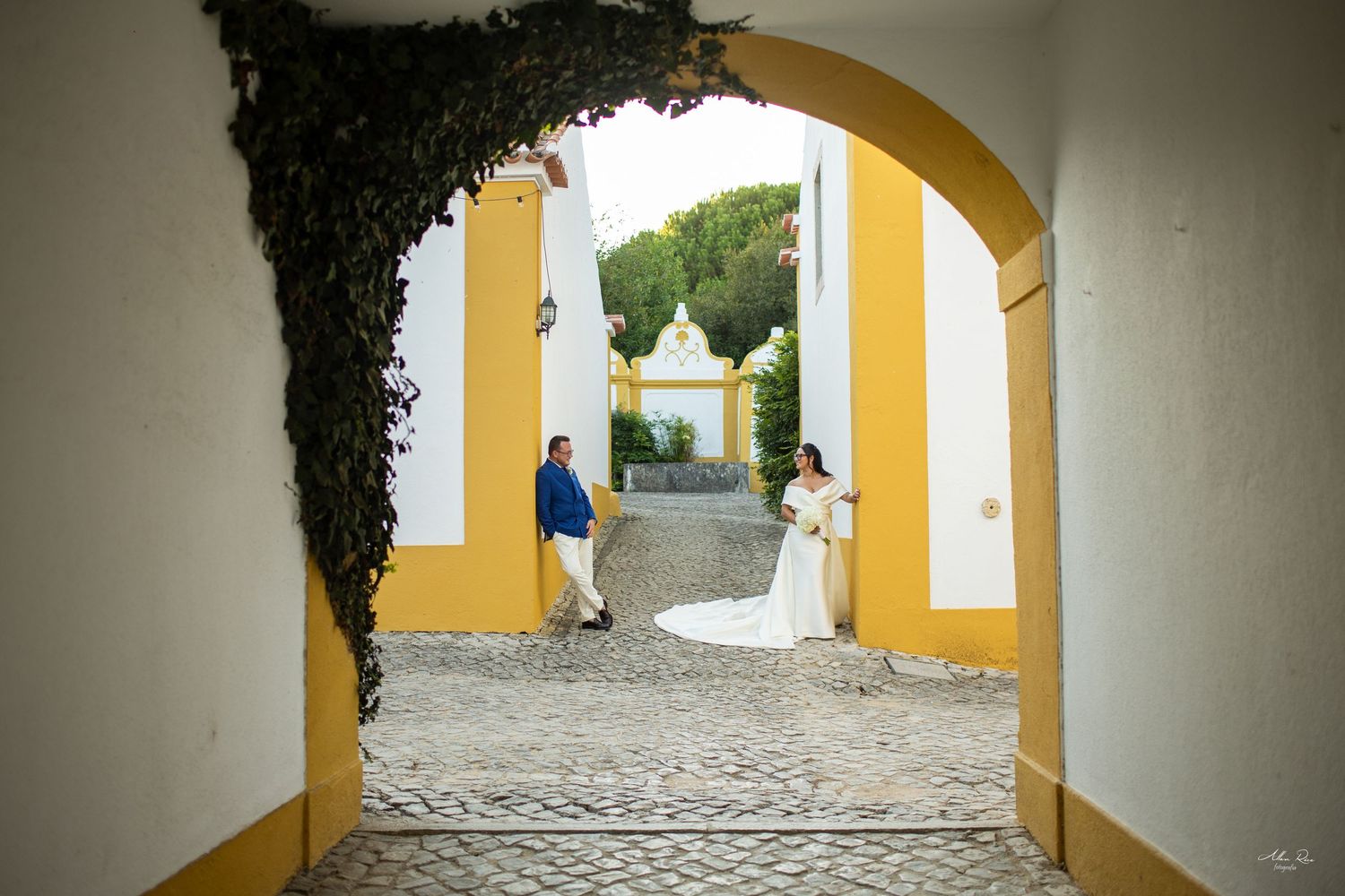 Bride and groom walk through yellow arched gateway on cobblestone path.