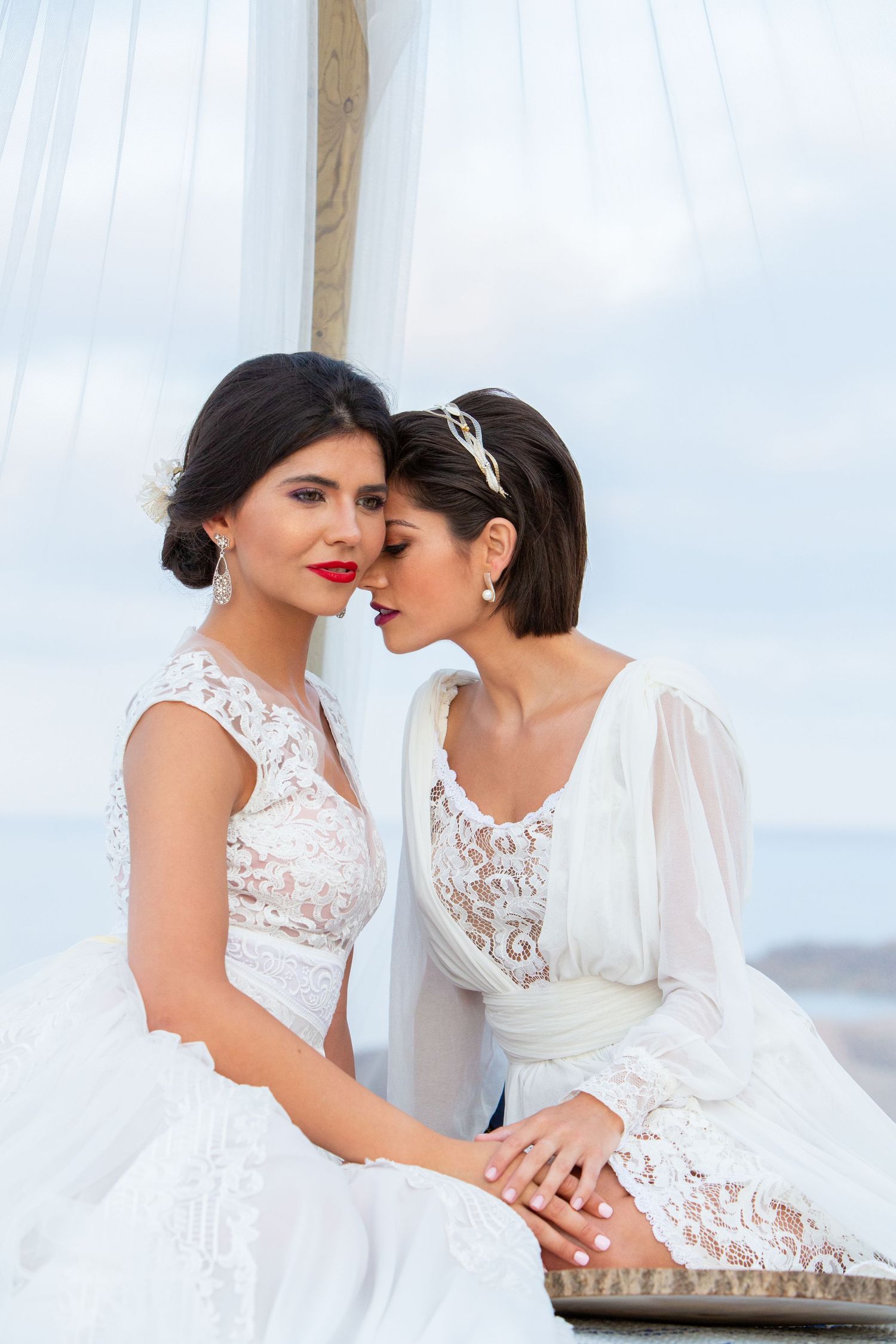 Two brides in white lace wedding dresses sharing an intimate moment by the sea under a canopy.