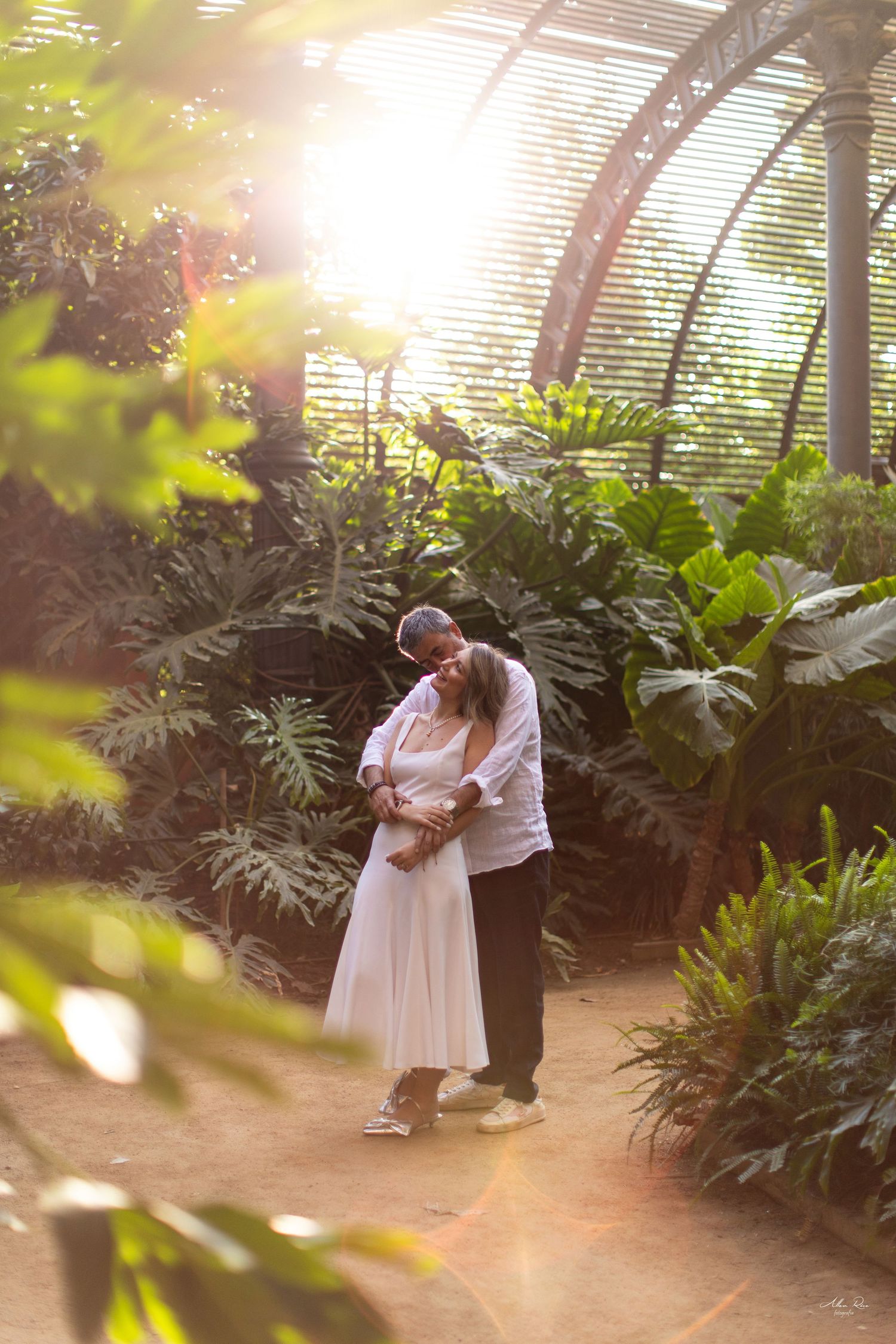 Couple sharing a tender embrace inside a sunlit greenhouse surrounded by lush tropical green plants.