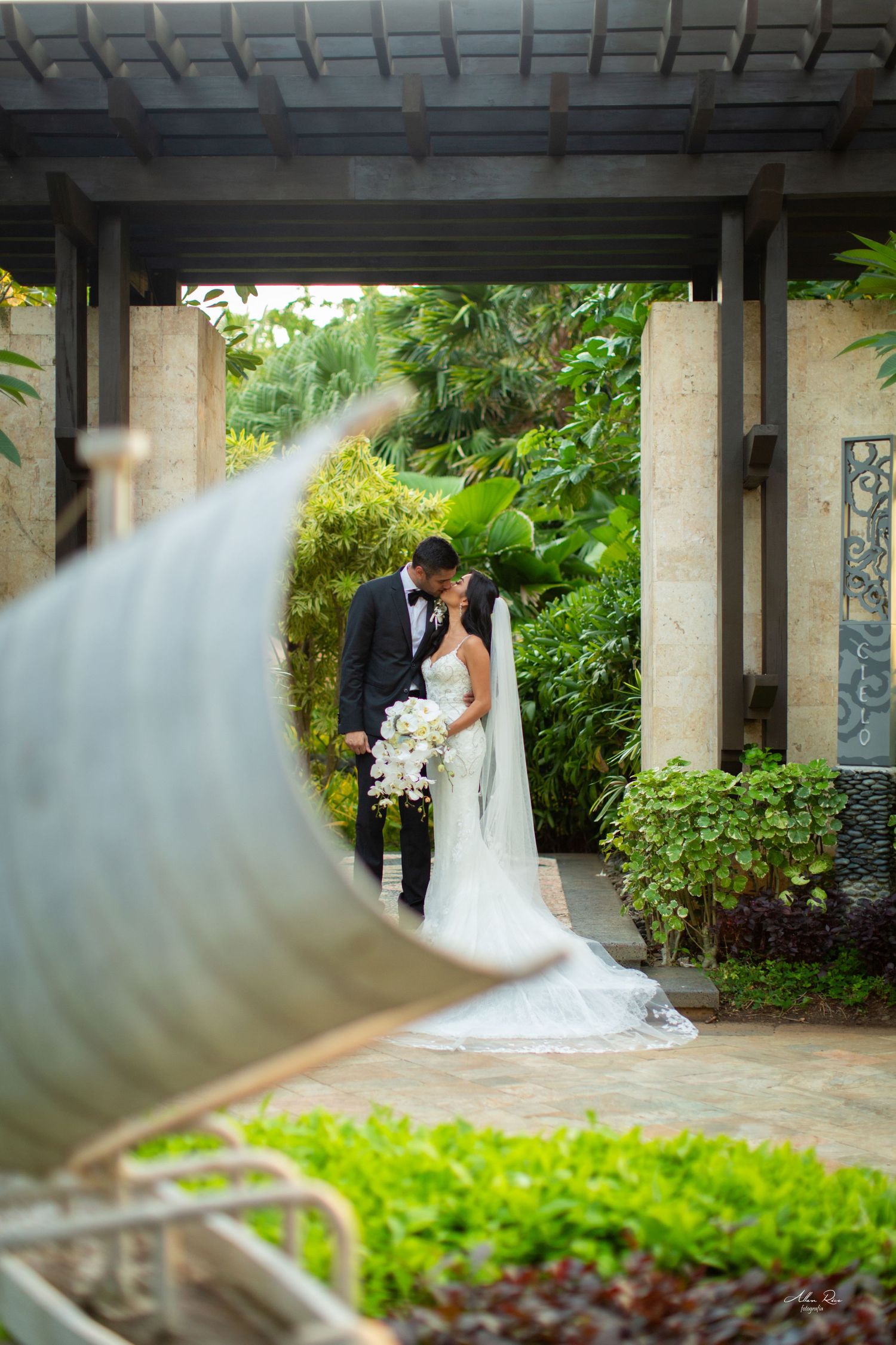 Bride and groom share a romantic kiss framed by a circular sculpture in a tropical garden pergola.