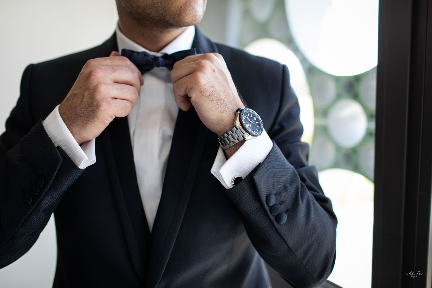 Groom adjusting black bow tie wearing a tuxedo with cufflinks and a watch.
