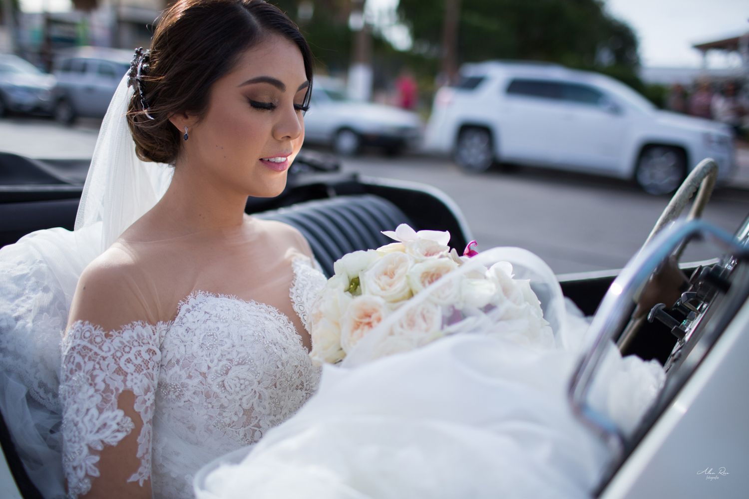 Radiant bride in lace off-shoulder gown holding bouquet while seated in vintage white car.