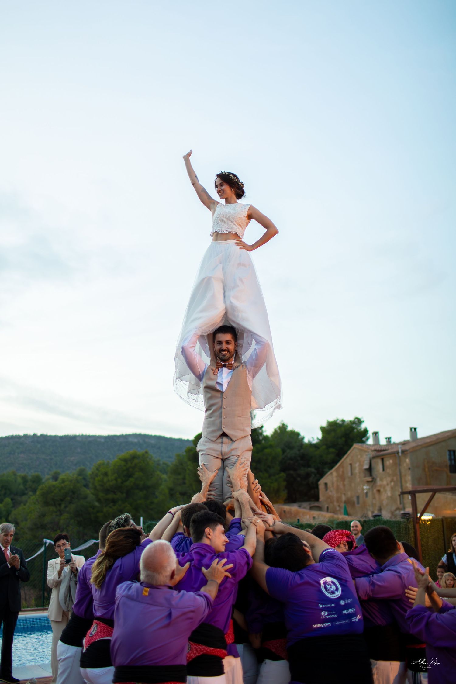 Bride triumphantly standing atop a human tower formed by wedding guests outdoors.