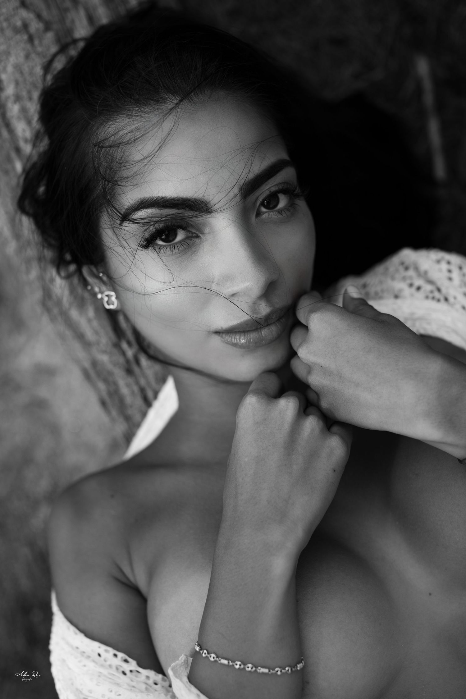 Black and white portrait of a young woman with elegant earrings and bracelet, gazing at camera with hands near face.