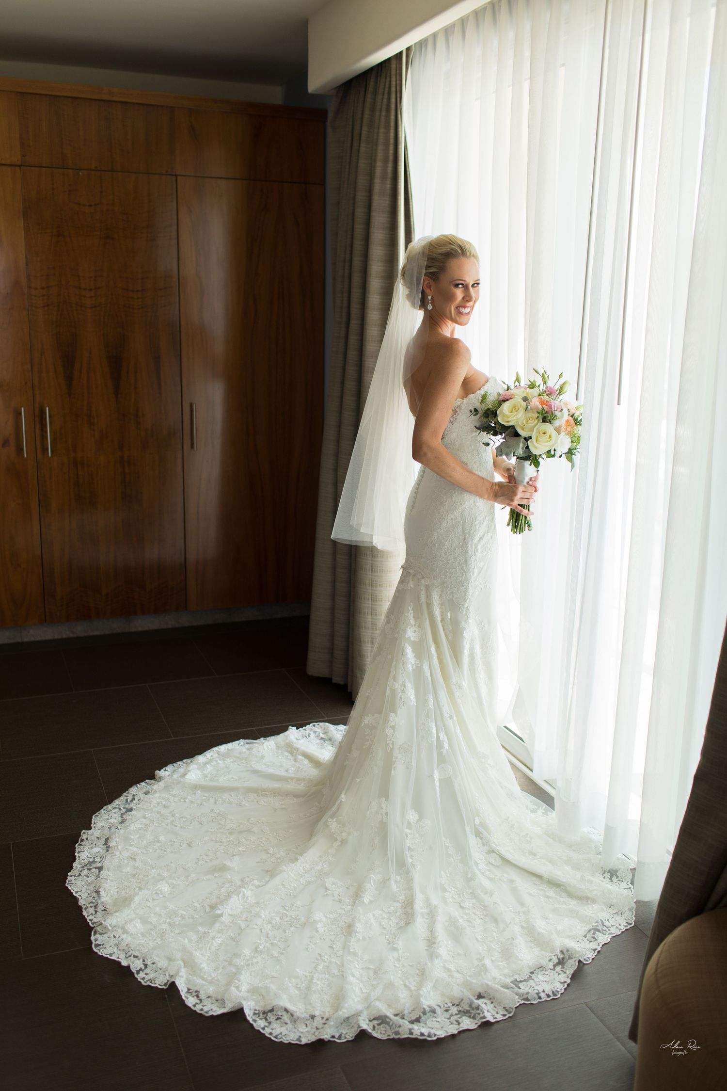 Elegant bride in a lace mermaid wedding dress with veil holding a bouquet by a bright window.