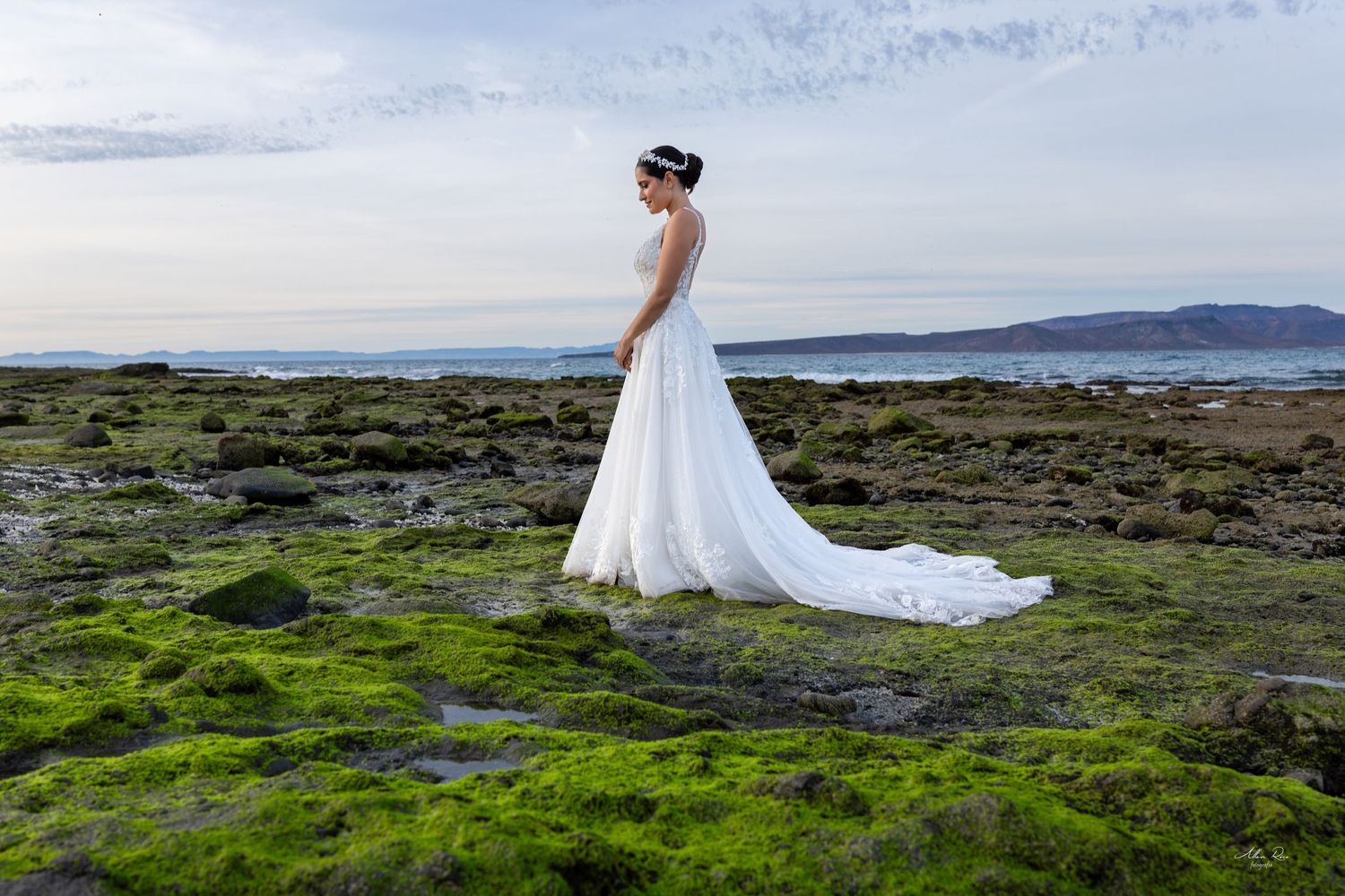 Elegant bride in flowing white gown with train stands alone on moss-covered rocky coastal landscape at dusk.