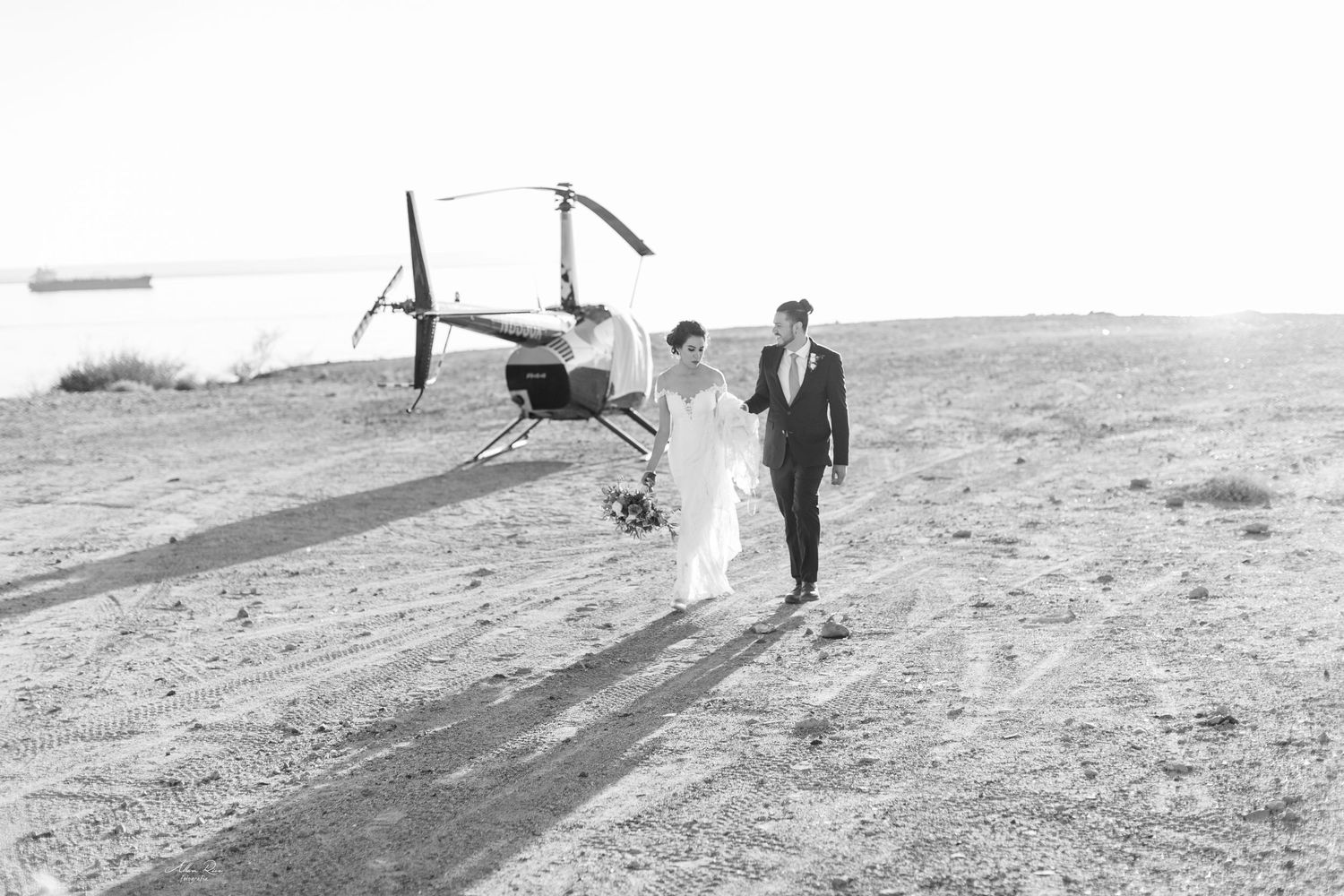 Bride and groom walk hand in hand near a helicopter on a scenic desert landscape in black and white.