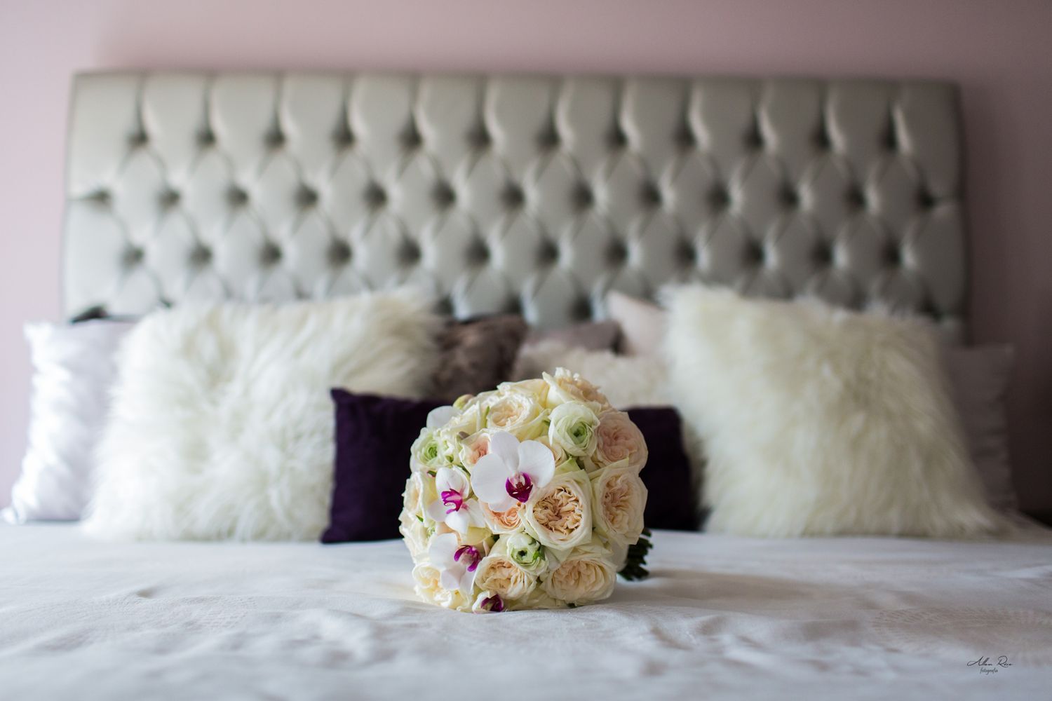 Beautiful white and pink bridal bouquet resting on bed with tufted headboard and pillows.