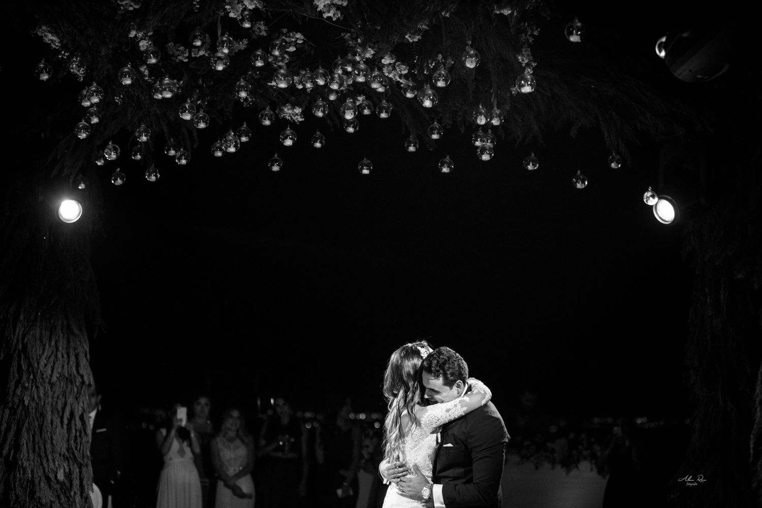 Bride and groom embracing during first dance under hanging lights in black and white.