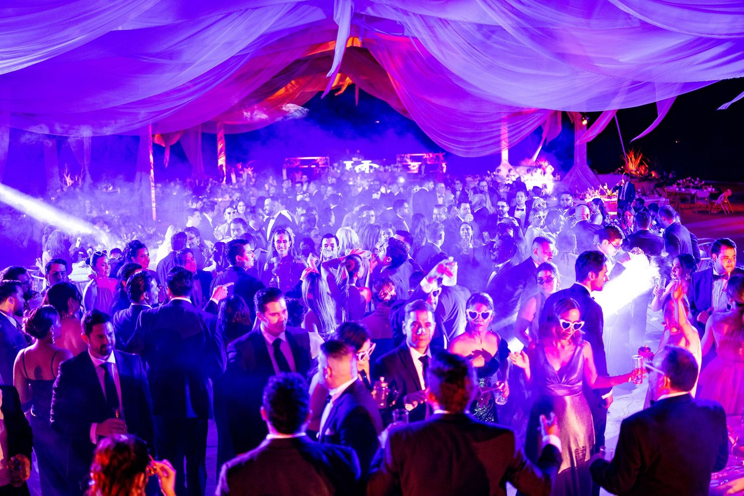 Elegant wedding reception dance floor packed with guests under purple and blue lights in a draped tent venue.