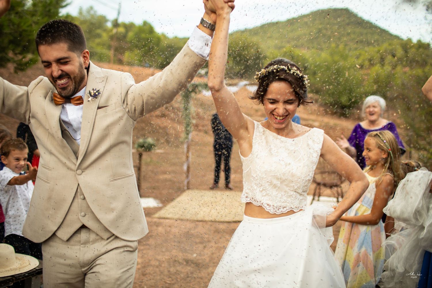 Newlyweds raise hands triumphantly as guests cheer during outdoor wedding celebration in sunshine.