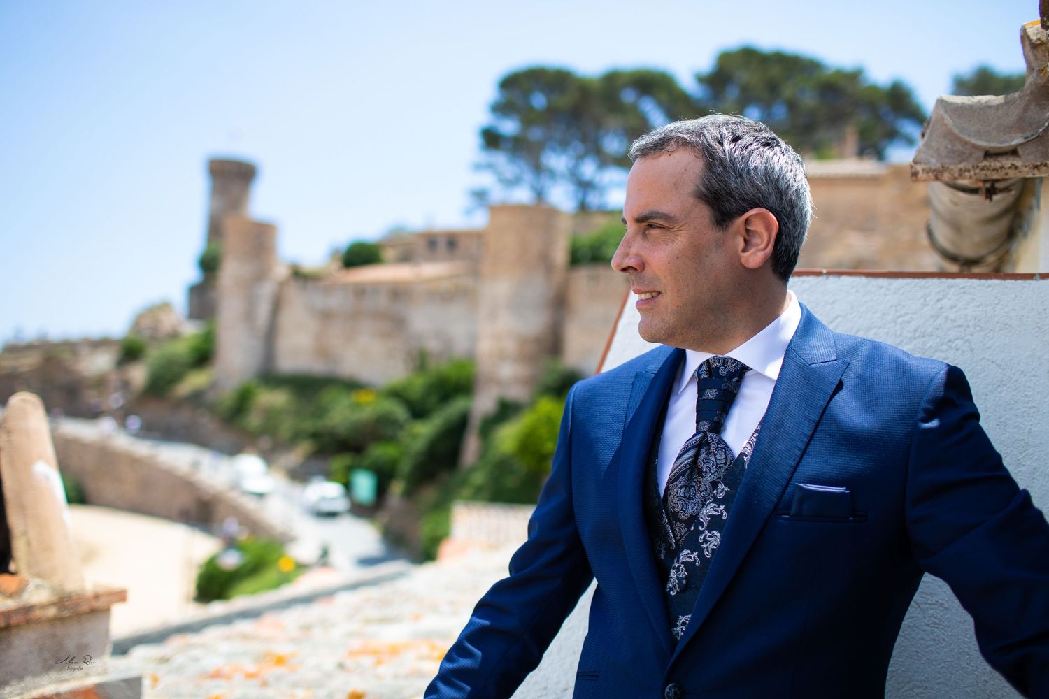Elegant groom in blue suit posing at a historic Mediterranean coastal castle.