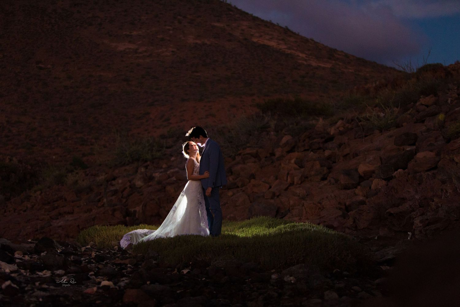Bride and groom share intimate moment on rocky hillside illuminated by dramatic nighttime portrait lighting.
