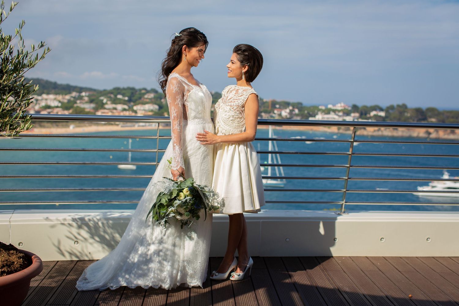 Two brides in white wedding dresses stand together on a seaside terrace with ocean views in the background.