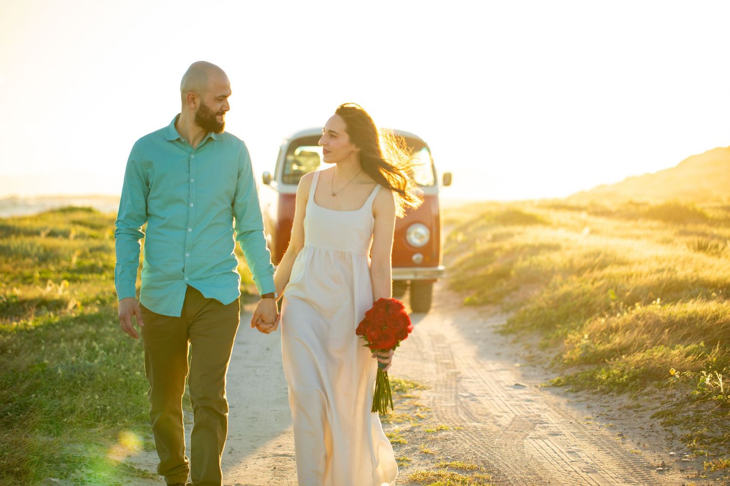 Smiling couple walking hand in hand at golden hour on a rural path with a vintage VW camper van behind.