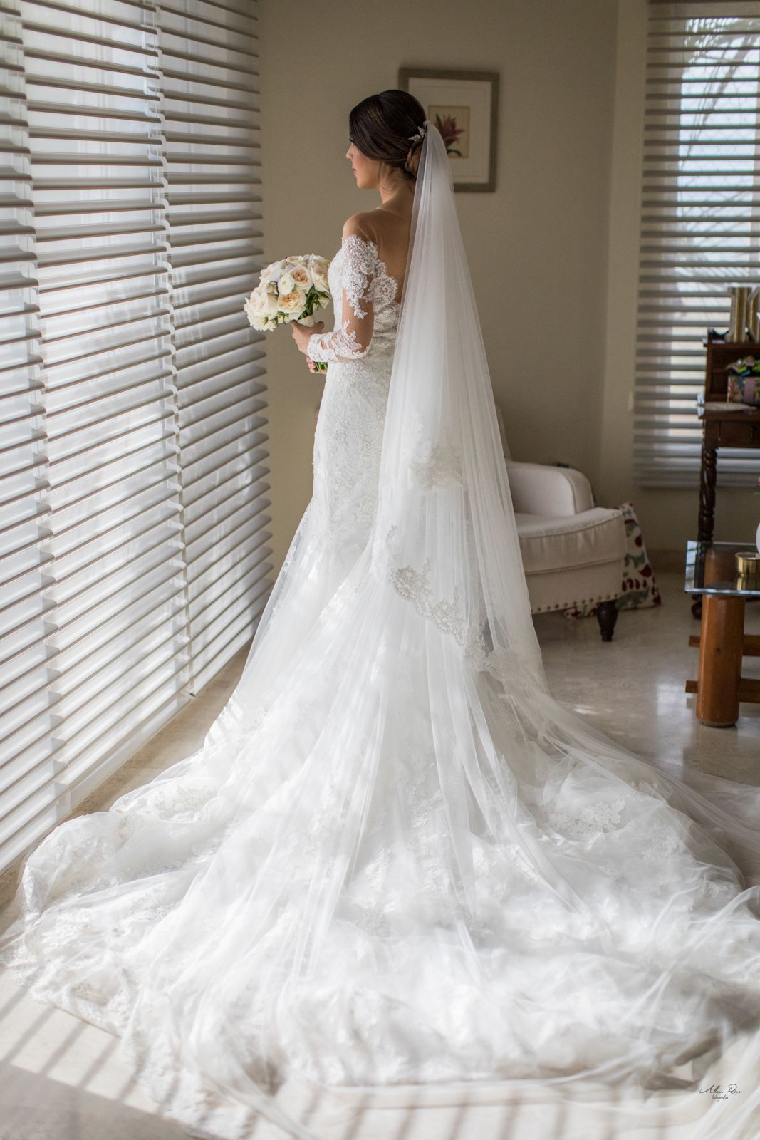 Elegant bride in lace wedding gown with long veil holding bouquet by window.