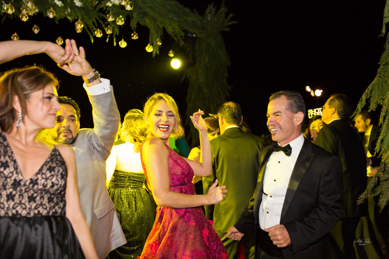 Guests in formal attire dancing joyfully outdoors at night under string lights at wedding celebration.