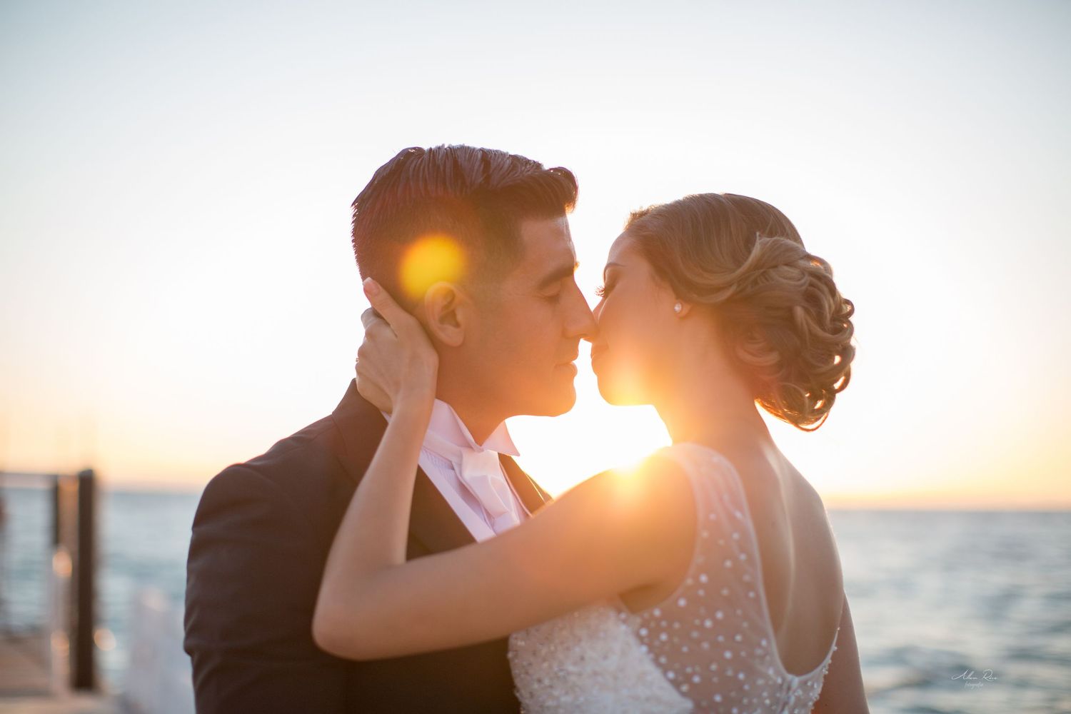 Bride and groom share a kiss at golden sunset by the sea in a romantic wedding portrait.