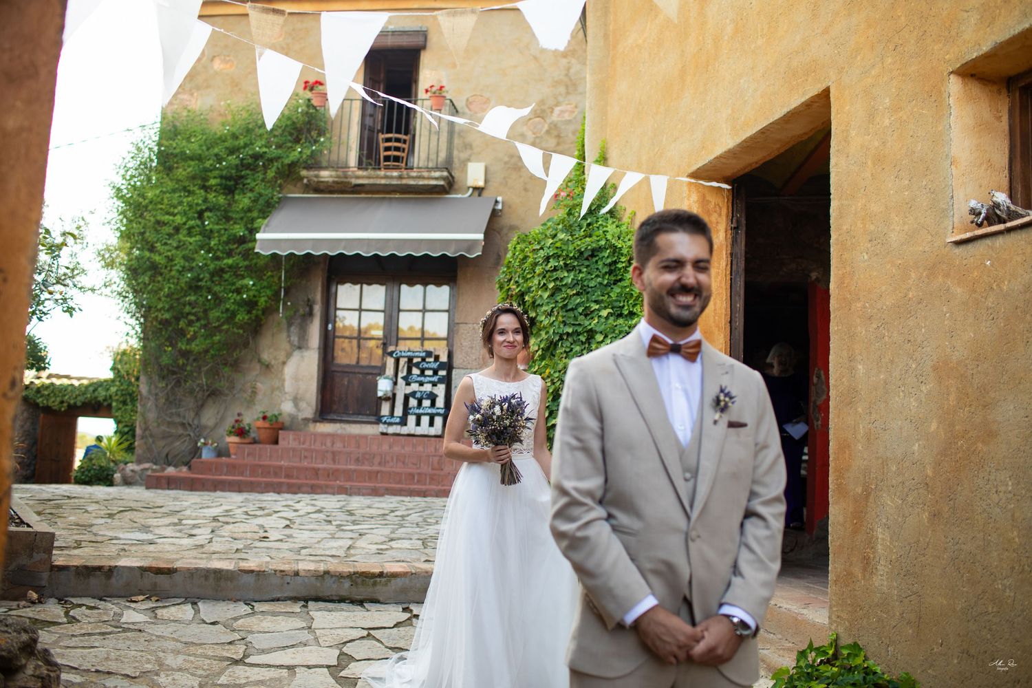 Groom in beige suit smiles as bride with bouquet stands behind in rustic courtyard.