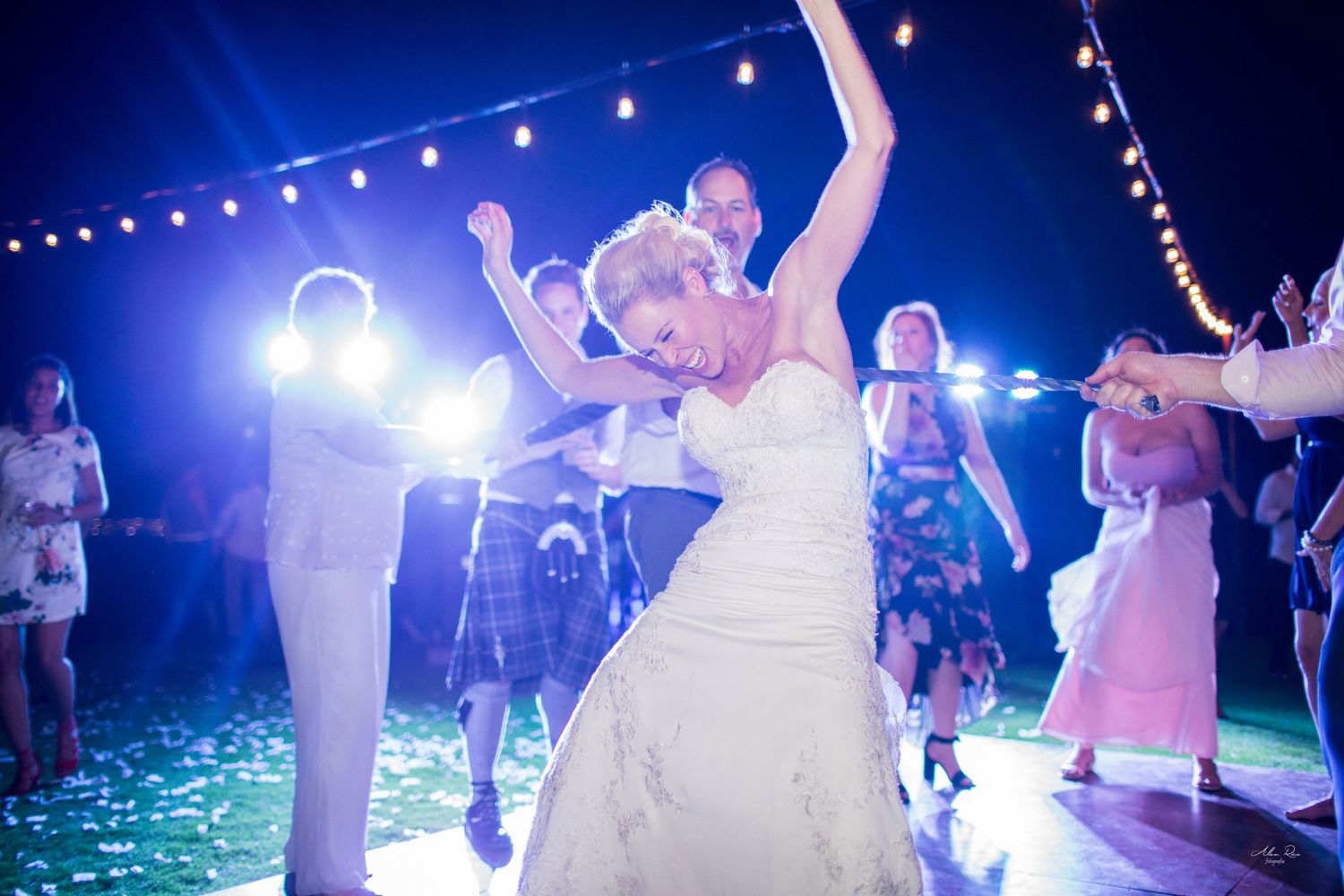 Bride in white ball gown dancing with arms raised at festive wedding reception under string lights.