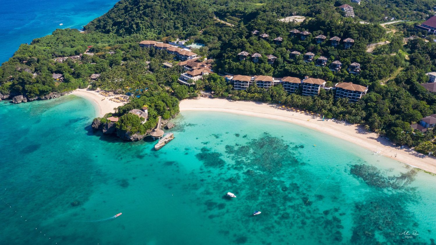 Aerial view of a tropical resort with turquoise waters, white sandy beach, and lush green hillside.