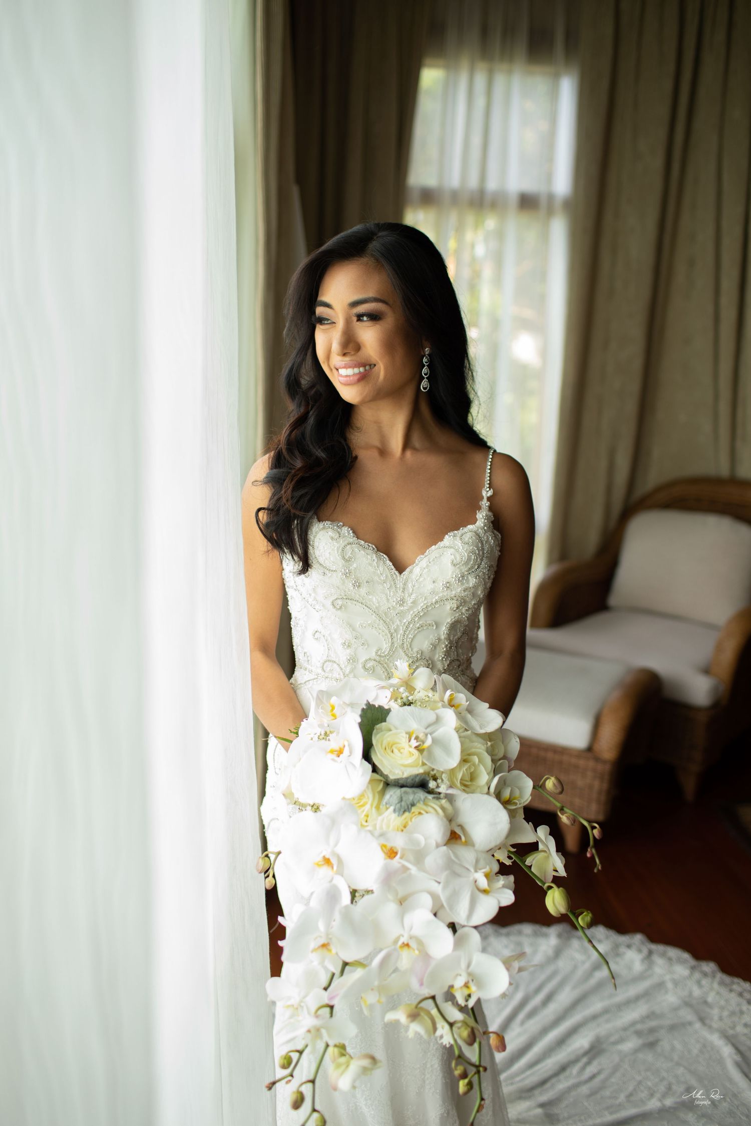 Radiant bride in lace mermaid gown smiling while holding cascading white orchid bouquet.