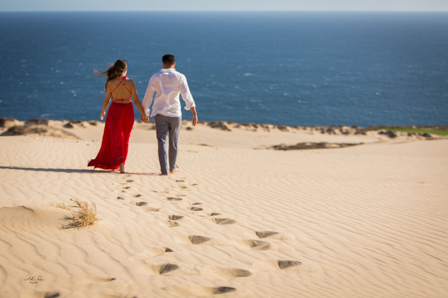 Couple walking hand in hand on sand dunes with ocean view, woman in red dress.