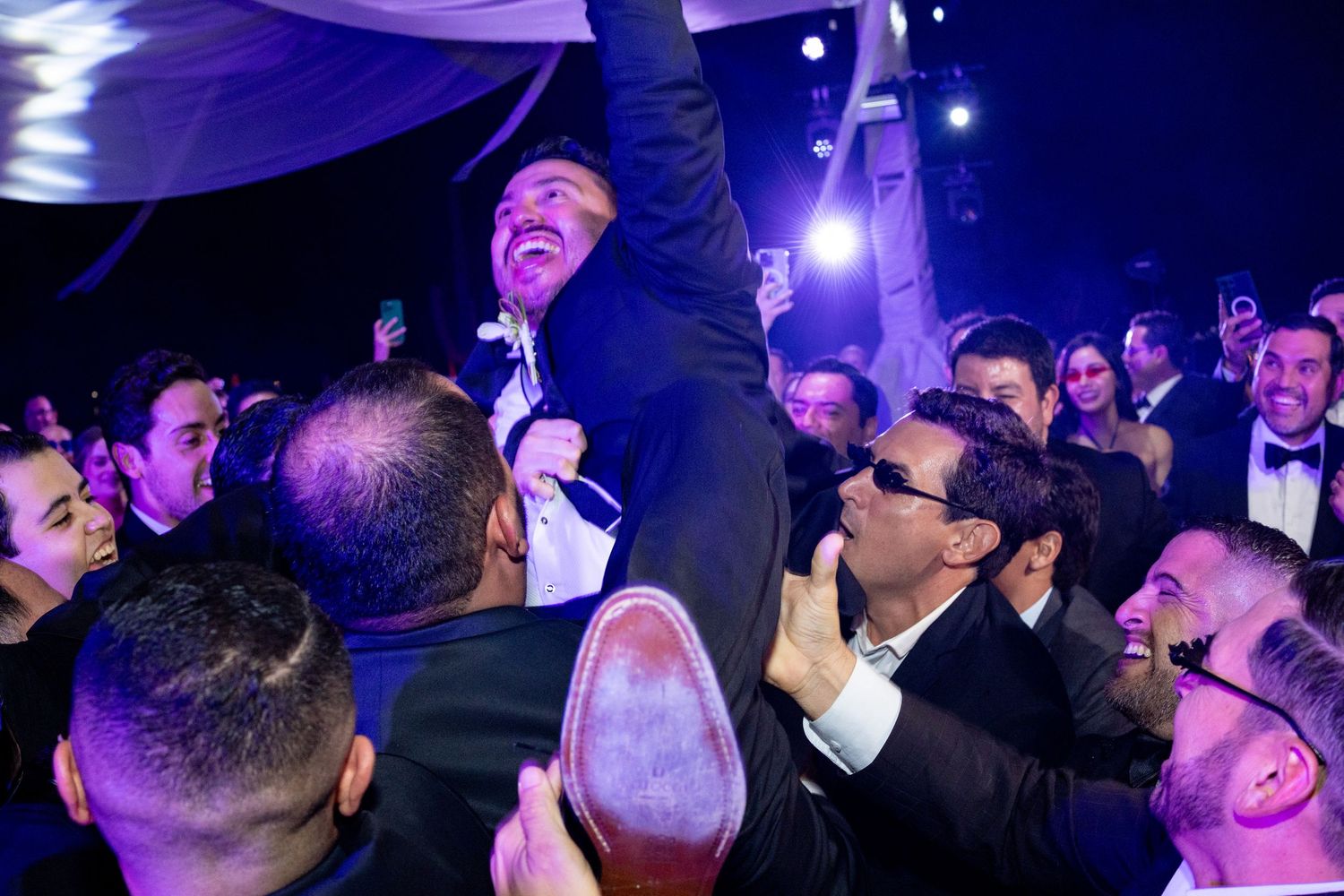 Groom lifted by celebrating guests in tuxedos on a lively, blue-lit wedding reception dance floor.