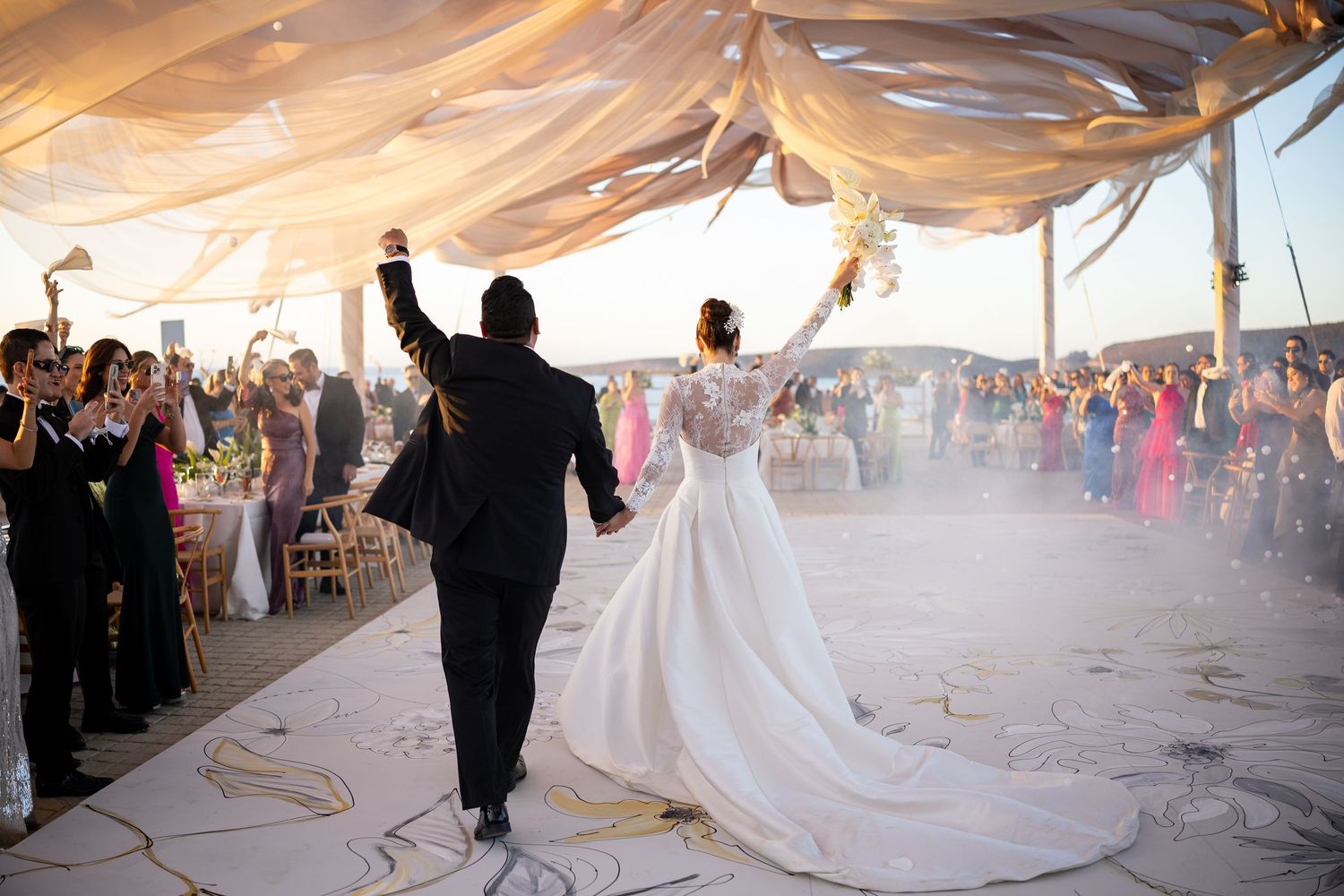 Bride and groom make grand entrance under flowing white canopy at elegant outdoor beach wedding reception.