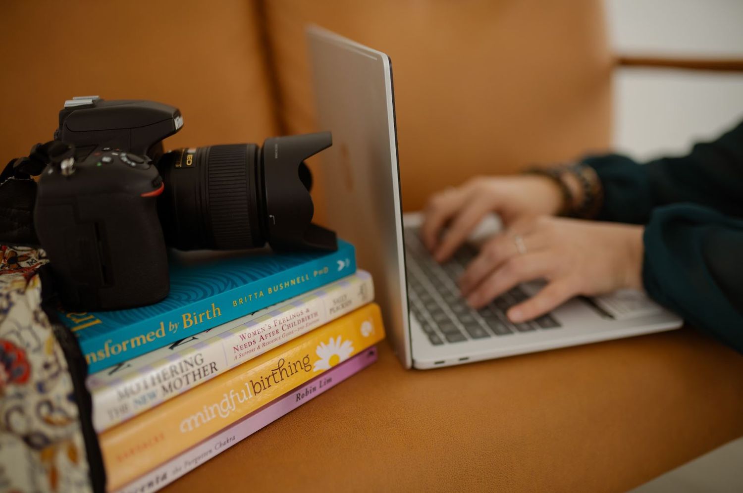 Person typing on laptop beside a stack of books topped with a DSLR camera on a leather couch.