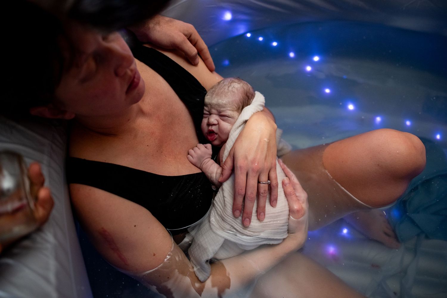 A newborn baby rests on mother's chest moments after a water birth, with blue lights in background.