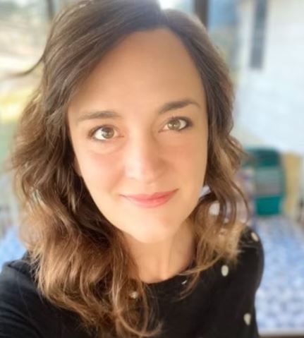 Smiling young woman with wavy brown hair and green eyes taking a selfie indoors in natural light.