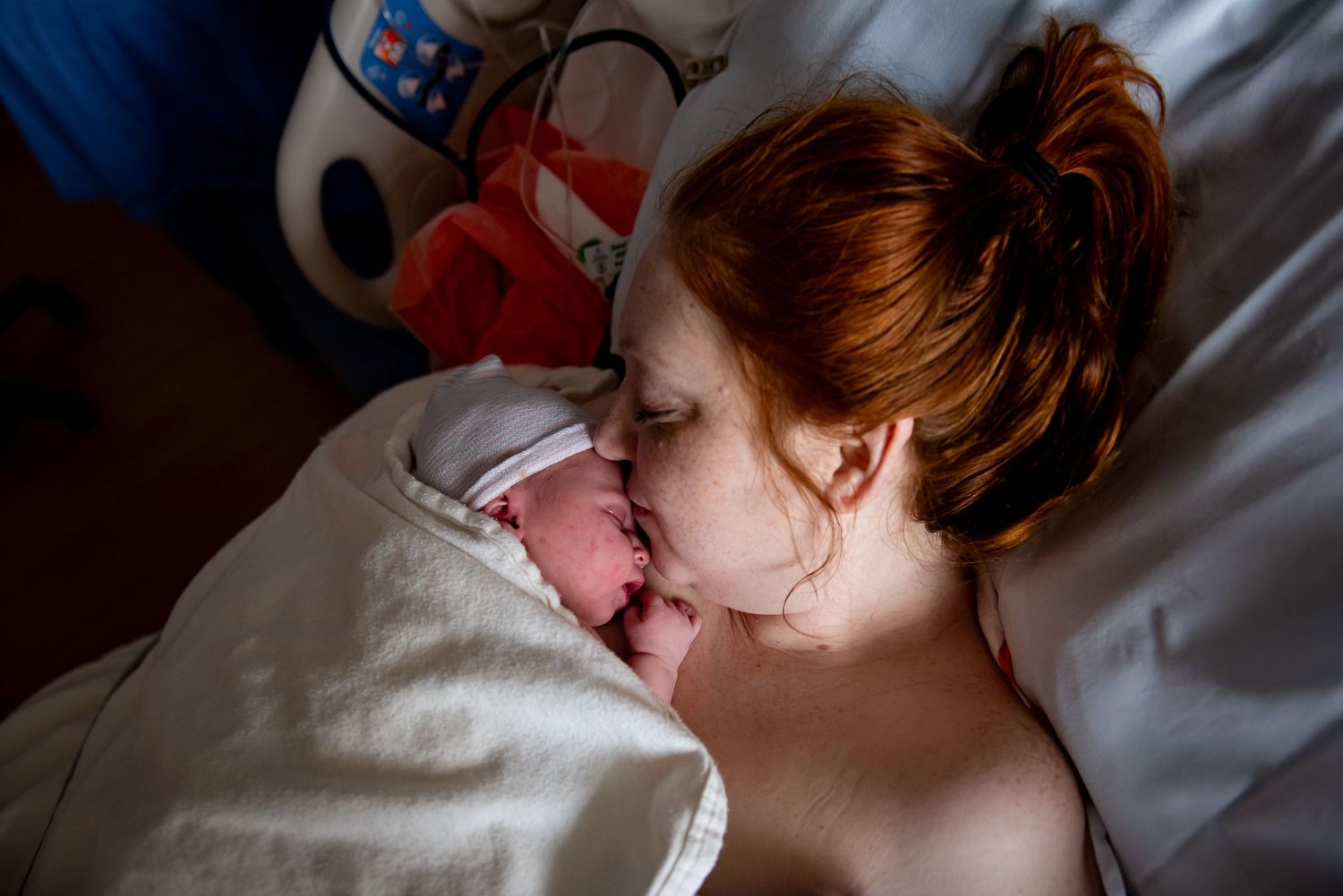 Red-haired mother gazes lovingly at her swaddled newborn in a dimly lit hospital room.