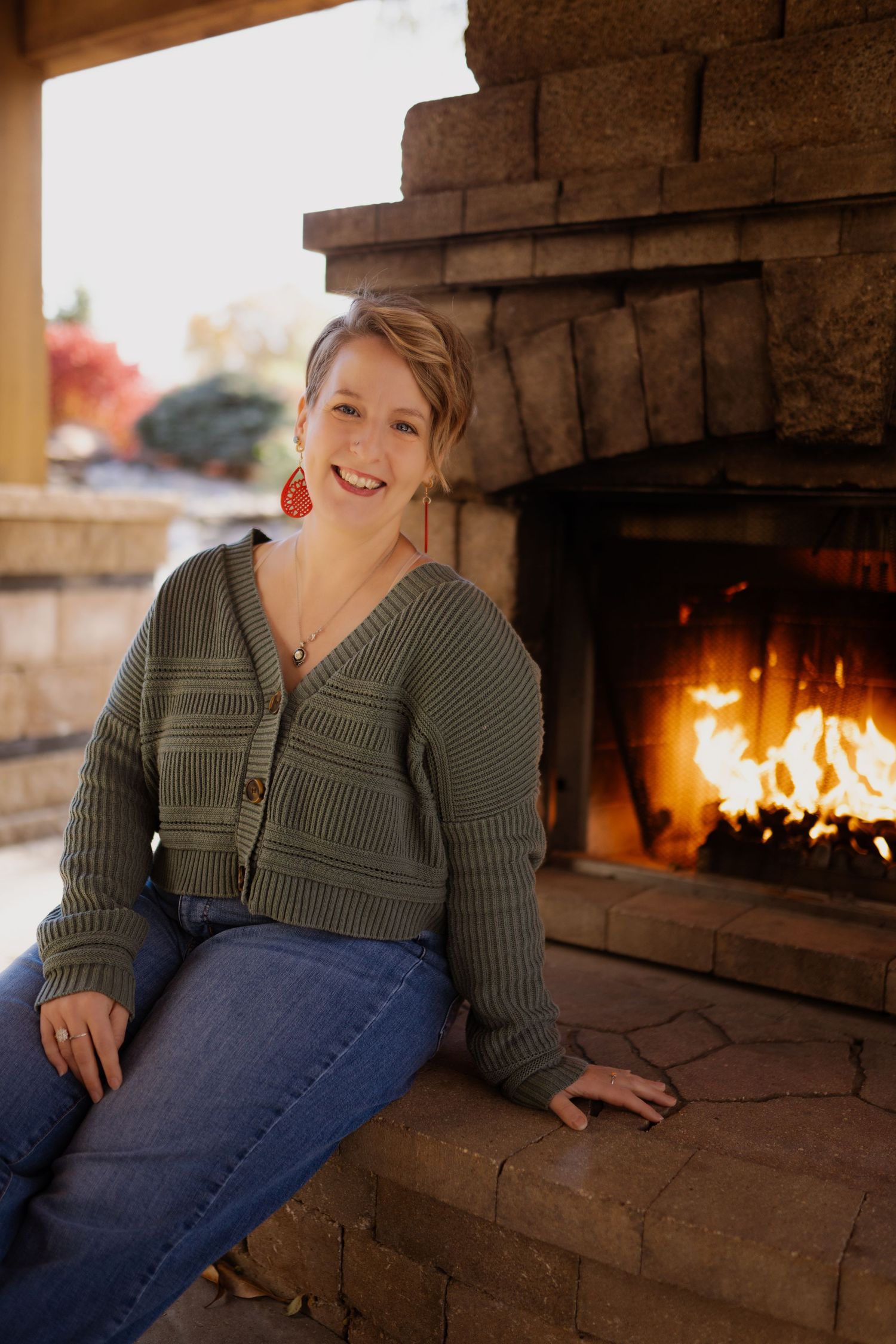 Smiling woman in green cardigan and jeans sitting by a stone outdoor fireplace on a warm autumn day.