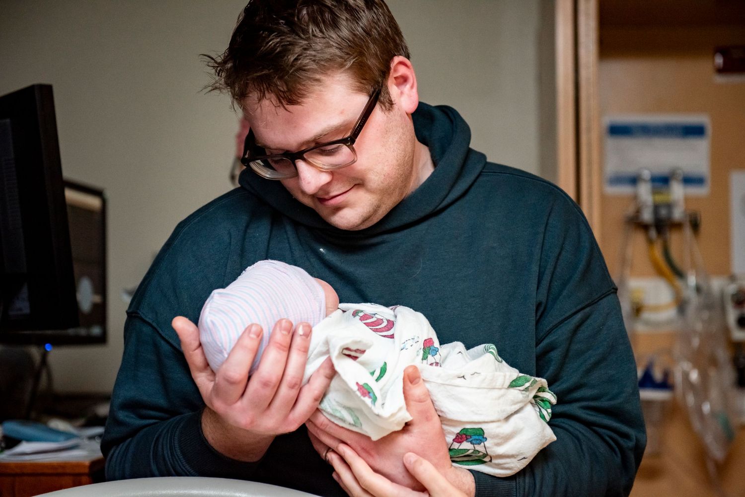 A young father in glasses tenderly holds his newborn baby in a hospital room.